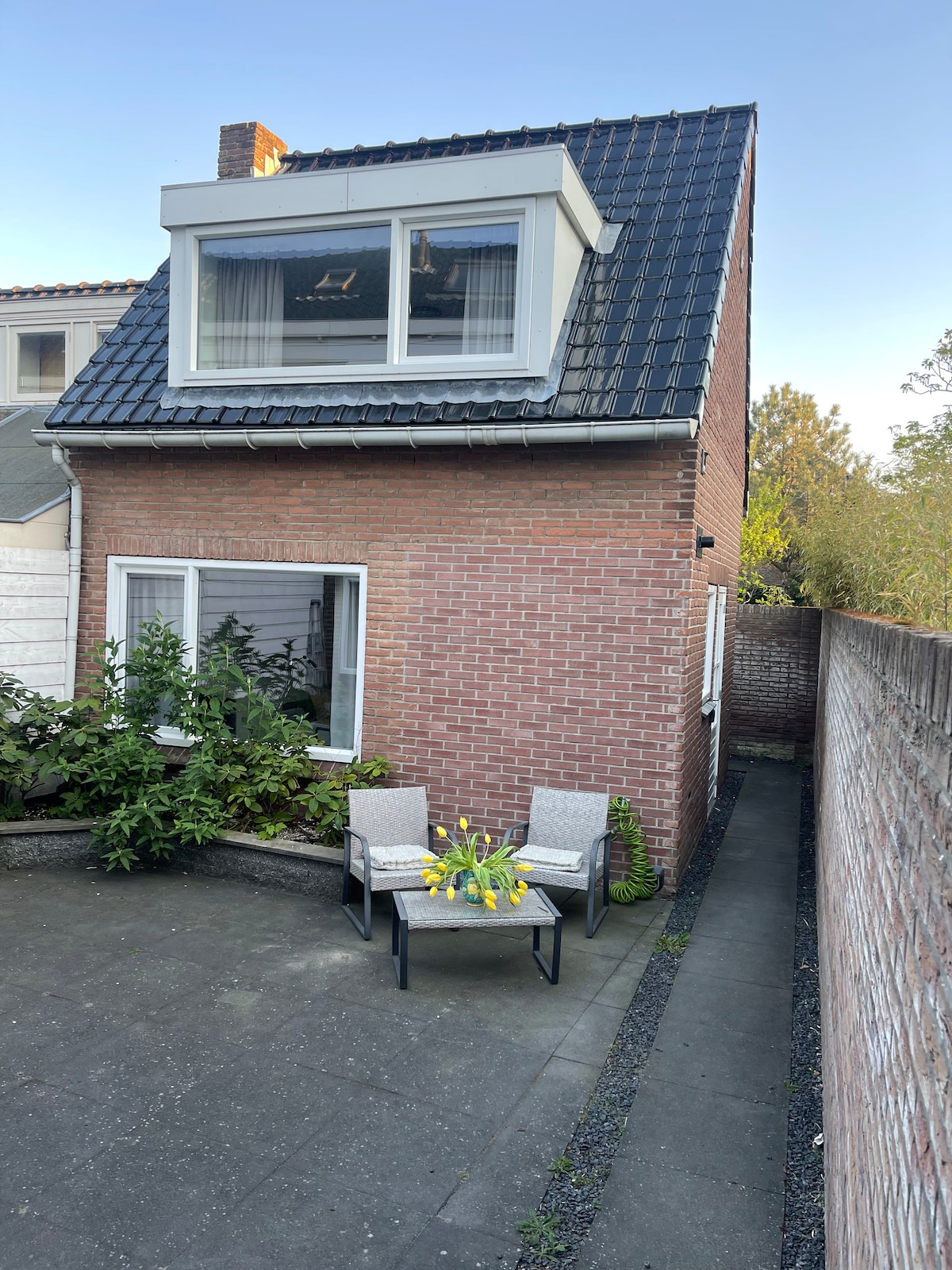 The exterior of the two-story house is shown, featuring a brick facade and a sloped roof. A small patio area is visible, furnished with two chairs and a table accented by a vase of yellow flowers. Lush green plants surround the patio, enhancing the outdoor space.