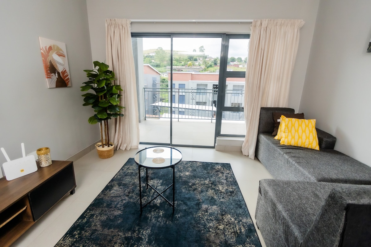 The open-plan living area features a comfortable gray sofa and a glass-top coffee table set on a textured area rug. Sliding glass doors lead to a private balcony, with sheer curtains framing the view and a potted plant adding a touch of greenery.
