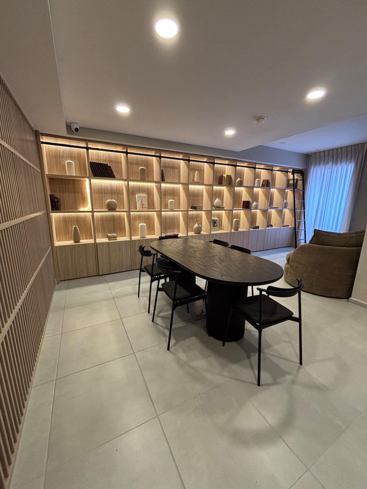 A spacious dining area is highlighted by a large, oval table surrounded by black chairs. A wooden shelving unit with integrated lighting displays various decorative items. Soft, natural light fills the room, with a cozy armchair positioned nearby and large windows partially covered by light curtains.