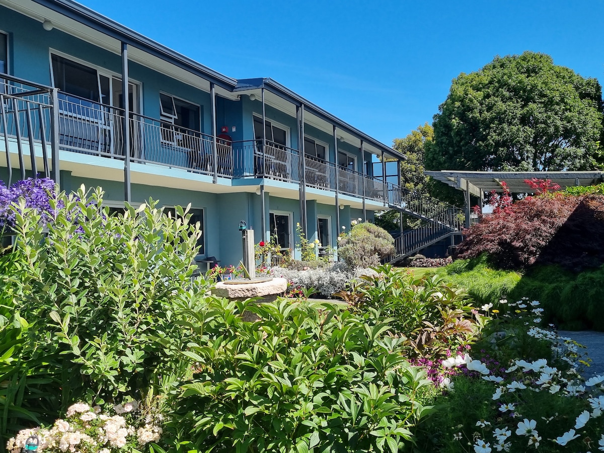 A multi-level building is surrounded by lush gardens filled with colorful flowers. Balconies are seen on each level, providing outdoor space for guests. The clear blue sky creates a bright atmosphere, and a variety of greenery enhances the tranquil setting.