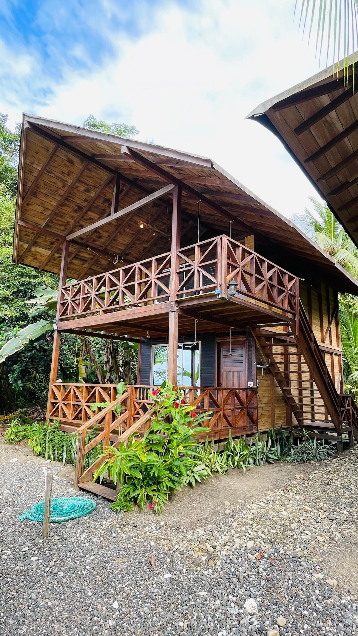 A wooden cabin is set amidst lush greenery, featuring a spacious balcony with decorative wooden railings. A staircase leads to the upper level, while vibrant plants surround the entrance. A calm pathway of gravel guides visitors to the inviting structure.