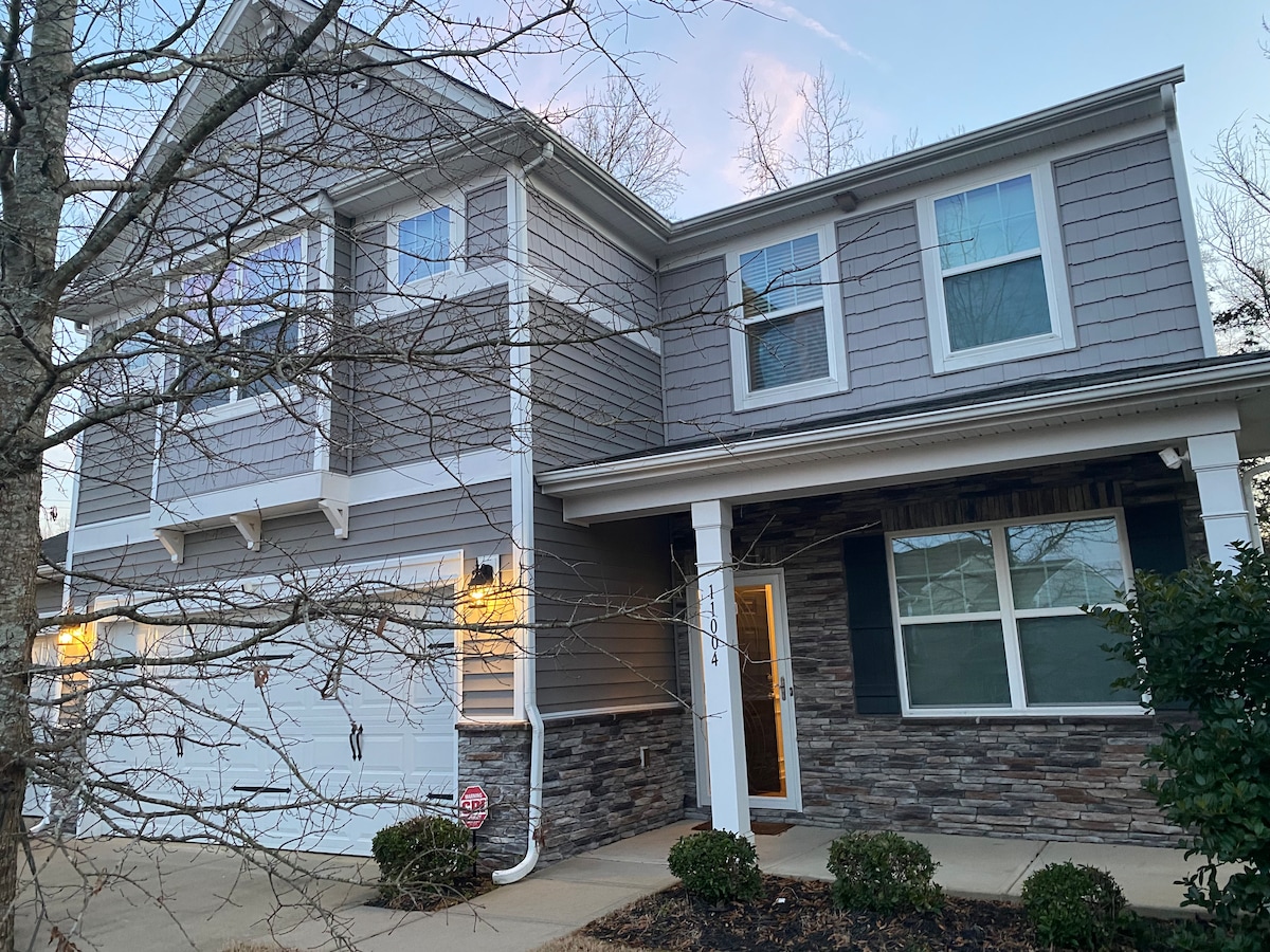 A charming exterior view of a two-story house is presented, featuring a combination of gray siding and stone accents. Large windows allow natural light to fill the inviting entryway, while landscaping includes neatly trimmed shrubs along the pathway.