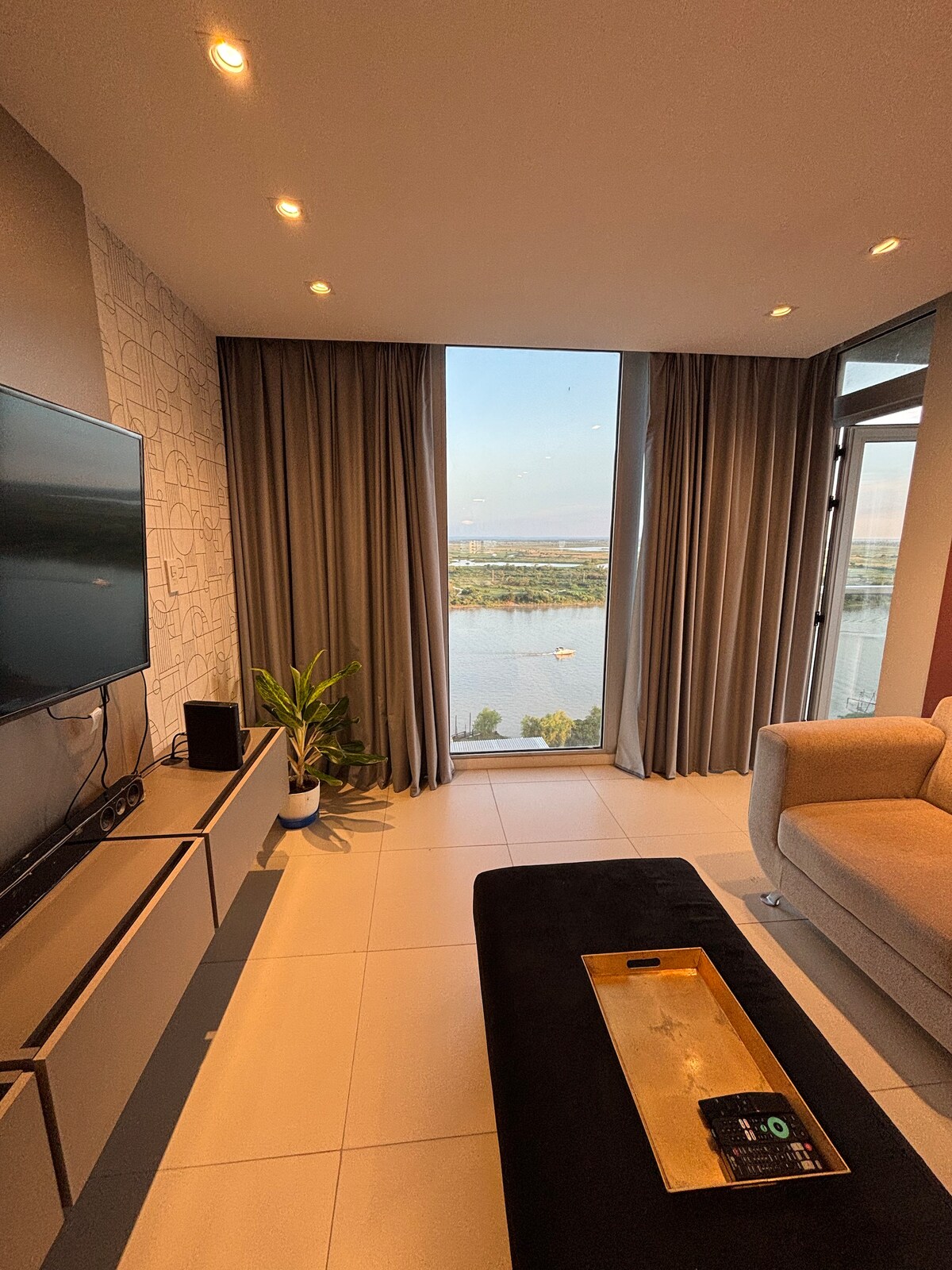 A living area features large windows offering views of the river and city. Soft curtains frame the glass, while a modern television is mounted on a wall unit. A potted plant adds a touch of greenery, complementing the neutral tones of the furnishings.