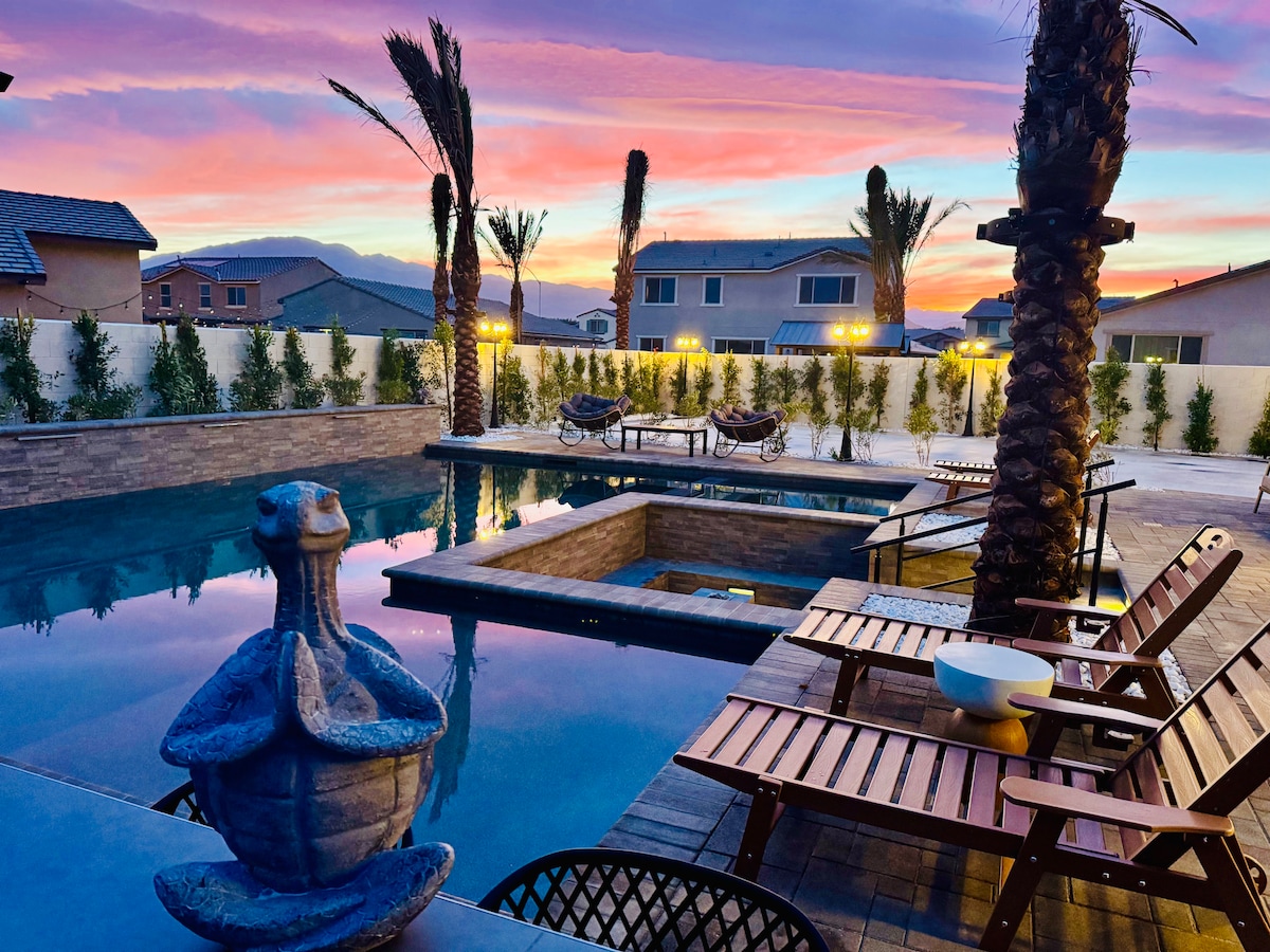 The outdoor pool area is framed by palm trees against a colorful sunset sky. A tranquil water feature sits prominently at the edge of the pool, while wooden loungers offer inviting spaces for relaxation. The spa is positioned nearby, surrounded by elegant stonework.