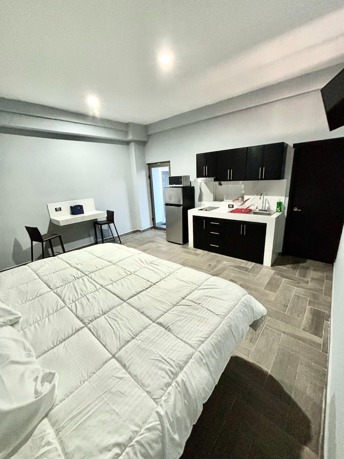 The spacious room features a large bed covered with a white quilt. A small kitchenette is equipped with dark cabinetry and a compact refrigerator. A dining table with two chairs occupies one side, while light enters through the door leading to a small outdoor area.