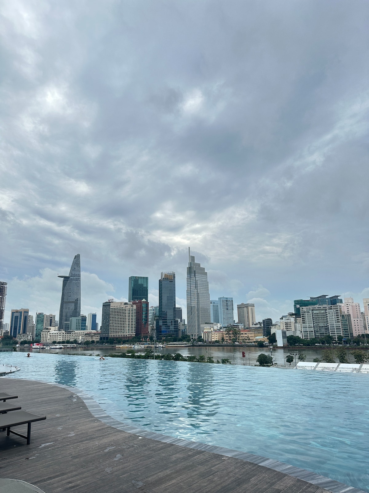An infinity pool is set against a backdrop of a city skyline, with modern skyscrapers reaching towards a cloudy sky. The water reflects the buildings while a wooden deck surrounds the pool, providing a space for relaxation.