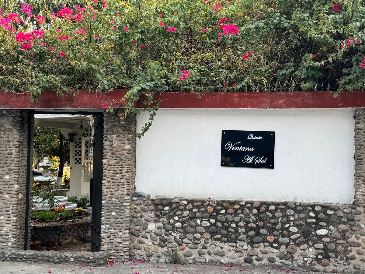 A decorative entrance is framed by a stone wall, showcasing a sign that reads 'Ventana Al Sol.' Vibrant blooms of bougainvillea cascade above, contrasting with the white surface. A glimpse of greenery may be seen through the open doorway, hinting at the serene surroundings.