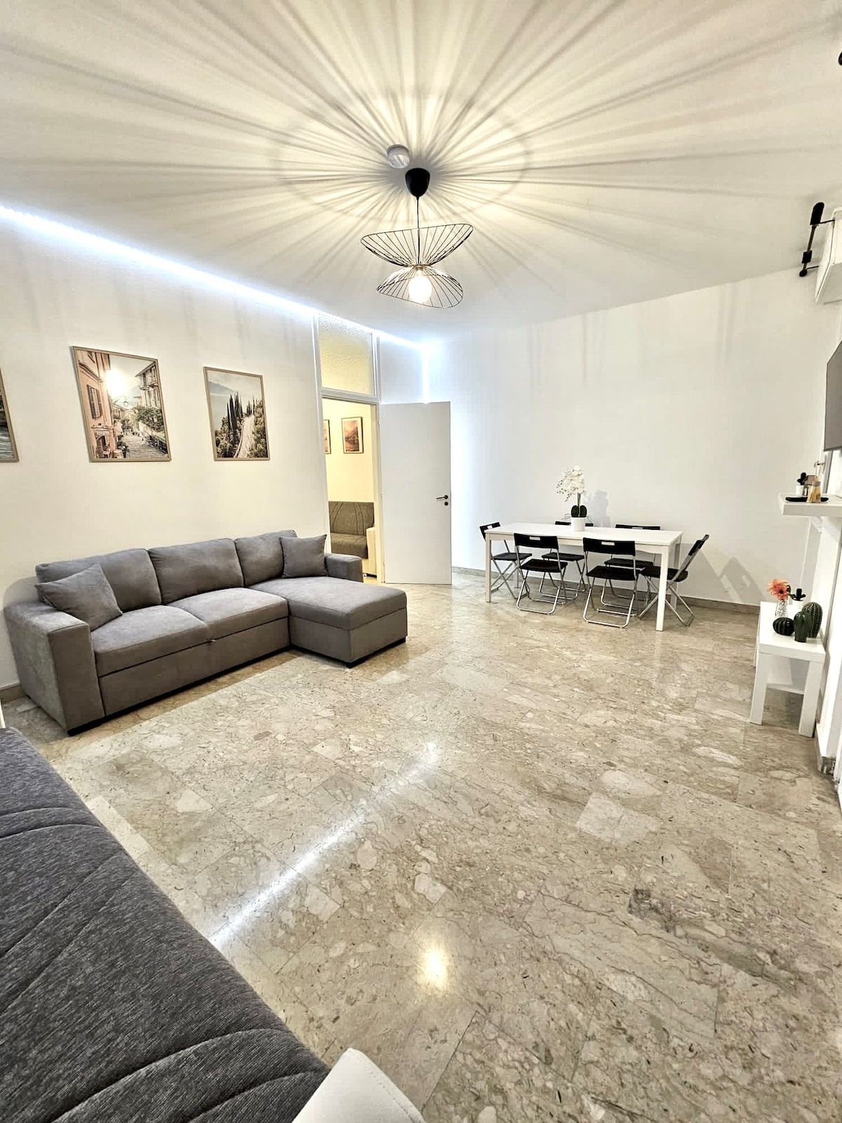A bright living area features a spacious gray sectional sofa, complemented by a modern dining table set for six. Natural light illuminates the room, highlighted by minimalist artwork on the walls. The polished stone flooring adds a touch of elegance to the space.