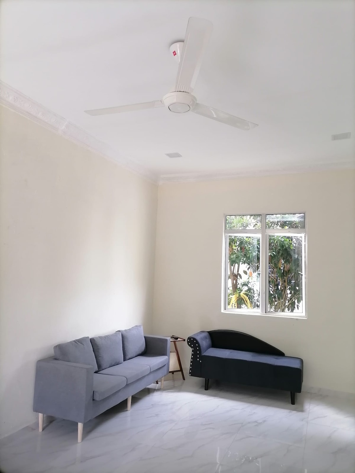 The image features a bright, airy living space with light-colored walls. A gray sofa is positioned adjacent to a dark blue chair. Natural light streams in through two windows, offering views of greenery outside. A ceiling fan is installed above, providing ventilation.