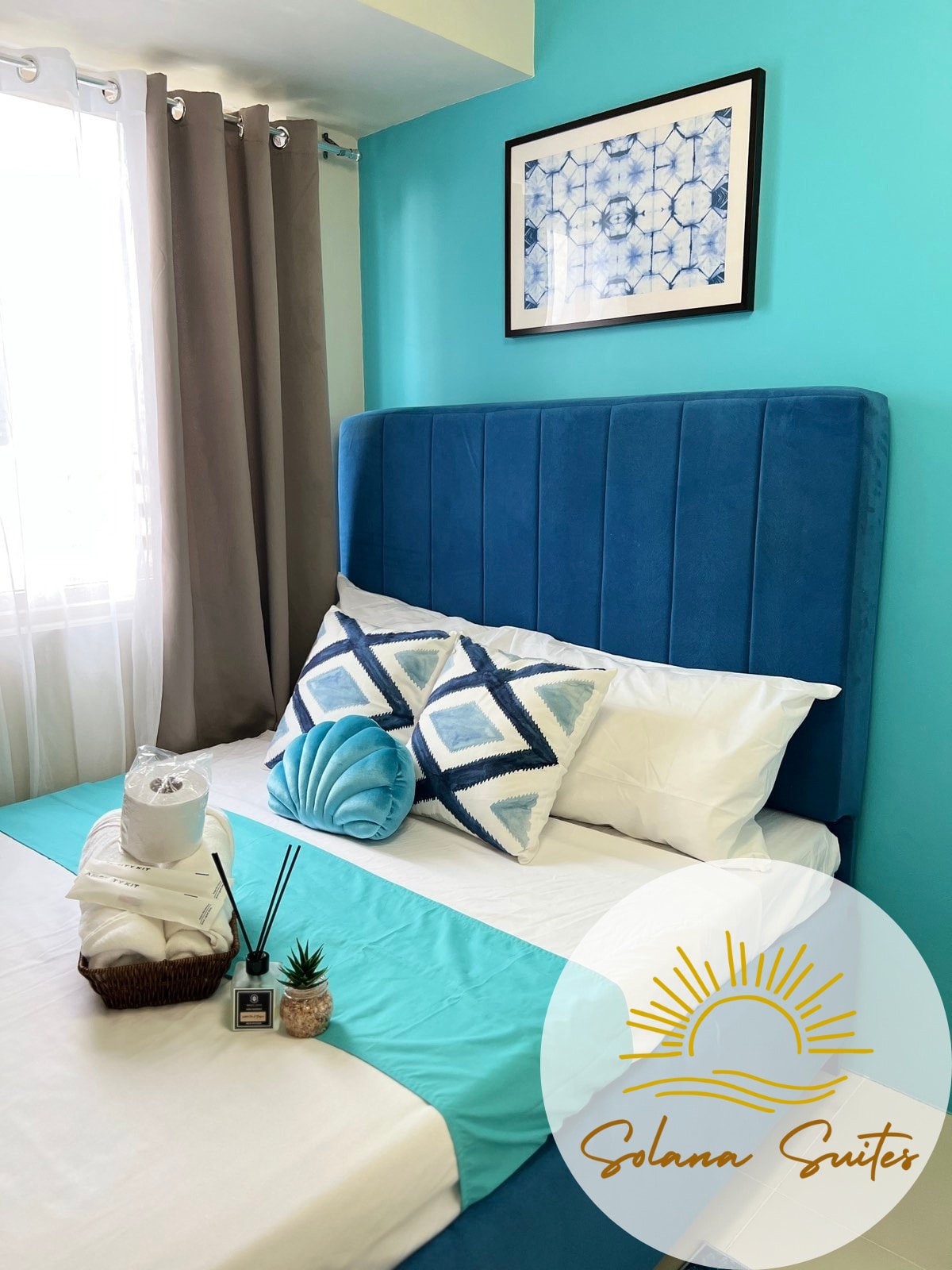 A tranquil bedroom features a spacious bed with a blue upholstered headboard and decorative pillows. Cool blue walls are complemented by soft, natural light filtering through sheer curtains. A neatly arranged basket with towels and a small plant adds a refreshing touch to the space.