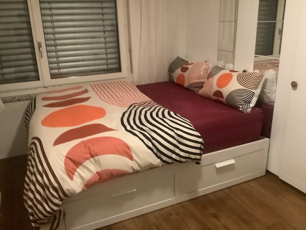 A cozy sleeping area is furnished with a spacious bed layered with a colorful duvet, featuring bold patterns in orange, purple, and black. Two decorative pillows complement the bedding. Natural light enters the room through two windows adorned with blinds, creating a pleasant atmosphere.
