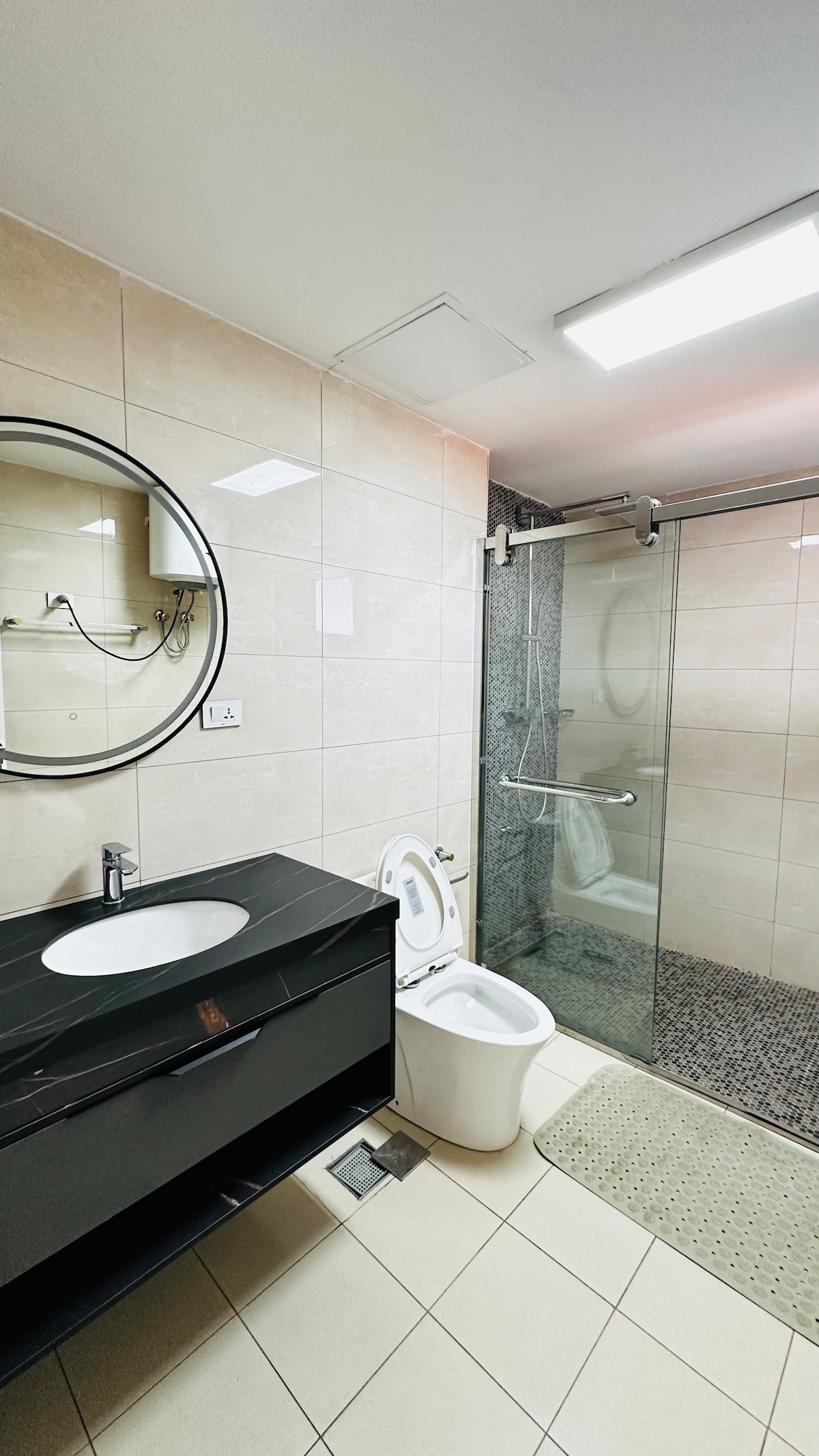 A modern bathroom features a sleek black vanity with a circular mirror above. A glass shower with a tiled enclosure is visible on one side, while a white toilet is neatly positioned beside it. Soft lighting enhances the clean, contemporary layout.