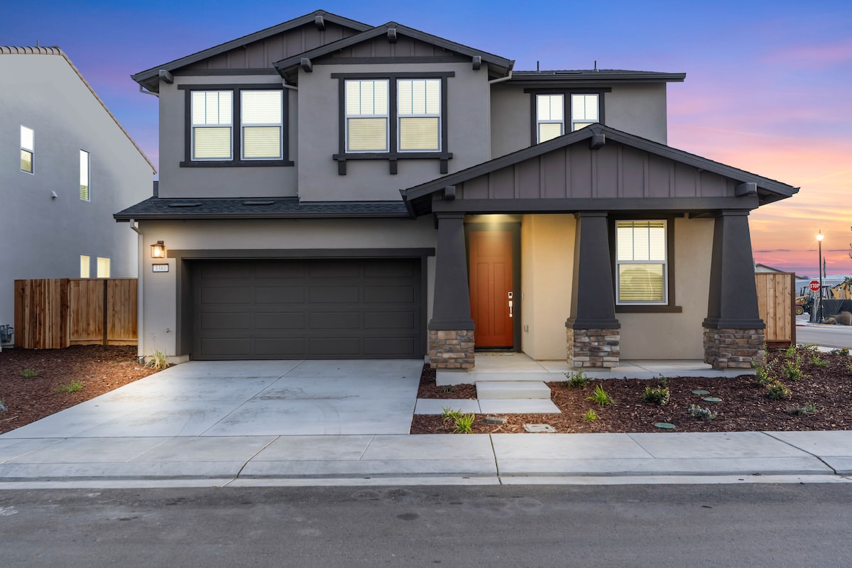 A modern two-story home features a stone-accented front porch and a well-defined entrance. Large windows illuminate the upper floor, complemented by a gray garage door. Surrounding landscaping includes freshly mulched areas, contributing to the contemporary curb appeal. The evening sky adds a warm hue to the exterior.