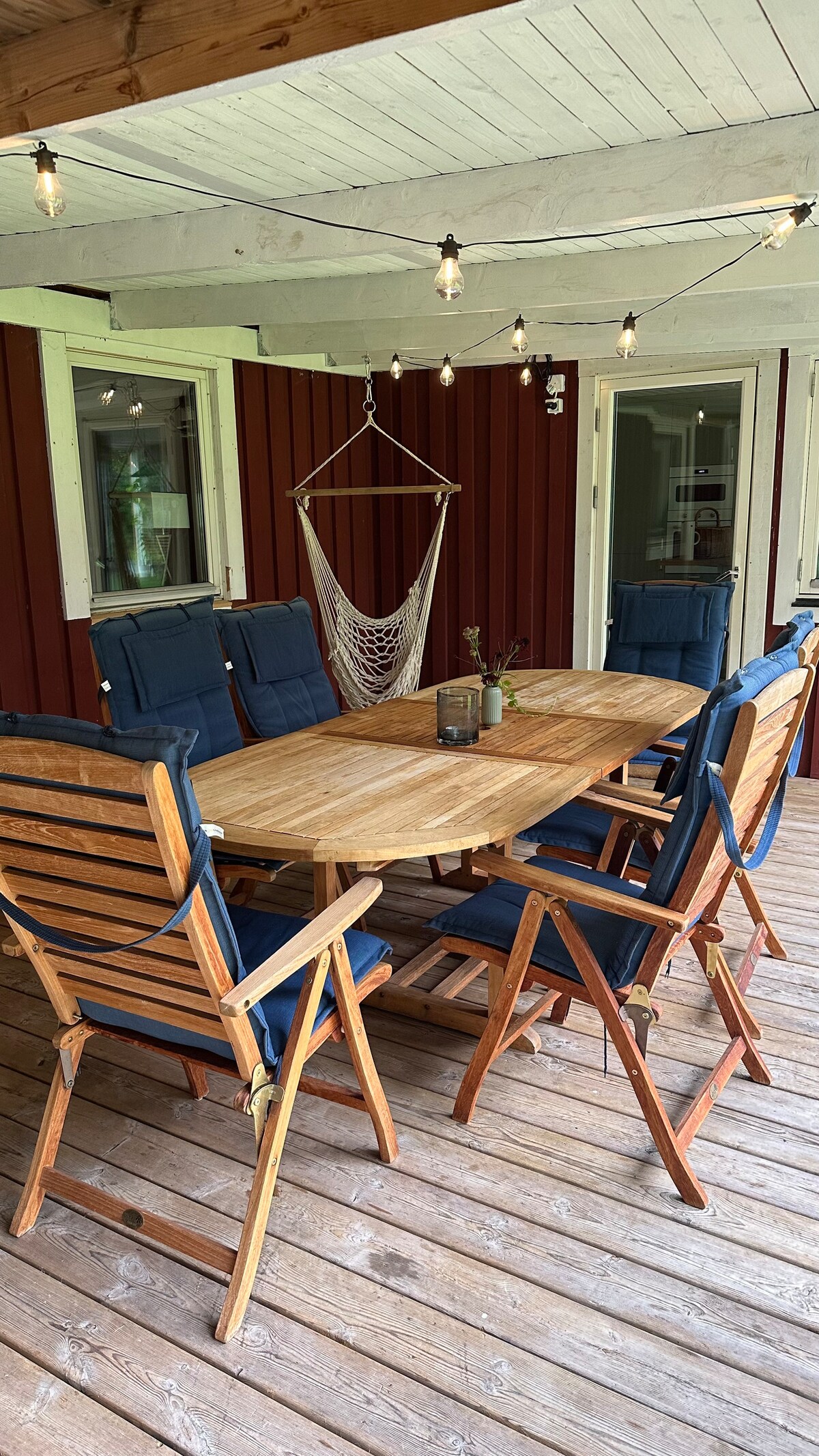 A wooden patio area features a large table surrounded by six folding chairs, all arranged on a wooden deck. Soft lighting from hanging bulbs creates a cozy atmosphere. A hammock is suspended nearby, and the red exterior walls of the house are visible in the background.