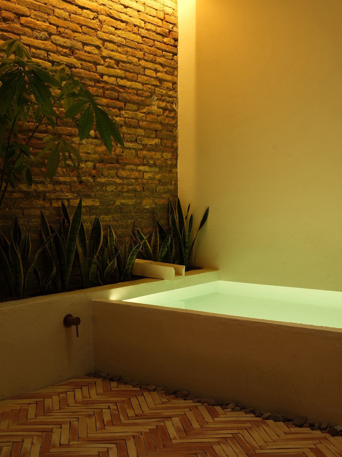 A serene spa area features a sleek soaking tub with softly glowing water. The surrounding space is enhanced by textured brick walls and lush greenery. Natural light is gently filtered, creating a calming ambiance for relaxation.