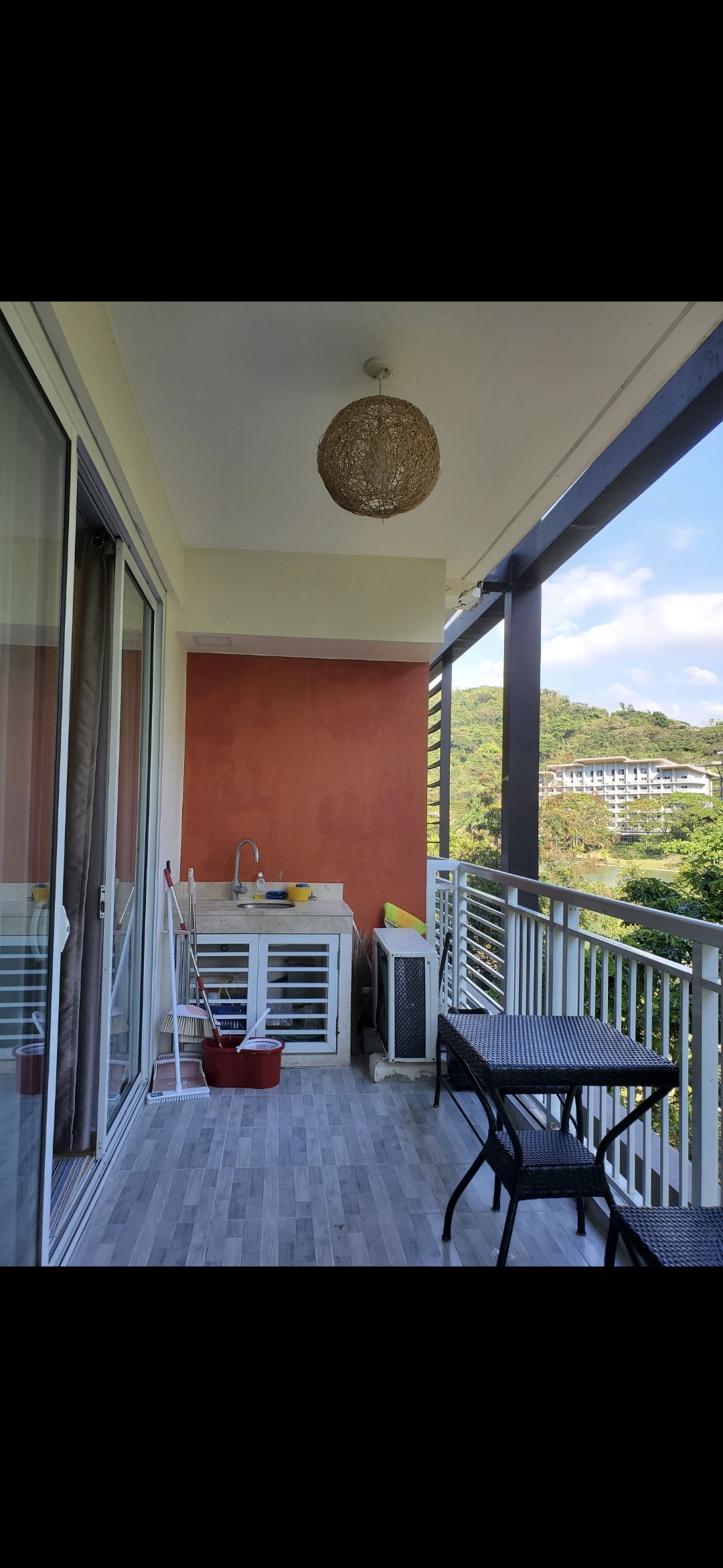 A balcony space equipped with a small outdoor sink and storage cabinet. Two black chairs and a matching table are positioned for relaxation. A decorative light fixture hangs above, and scenic views of surrounding greenery and nearby structures are visible in the background.