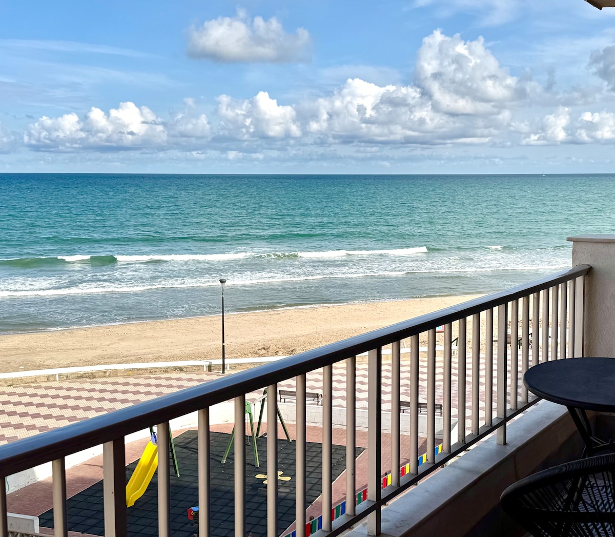 An expansive balcony overlooks a sandy beach and the vibrant blue sea, which reflects the partly cloudy sky. The scene captures gentle waves, while colorful playground equipment is visible along the beachfront, enhancing the beachside atmosphere.