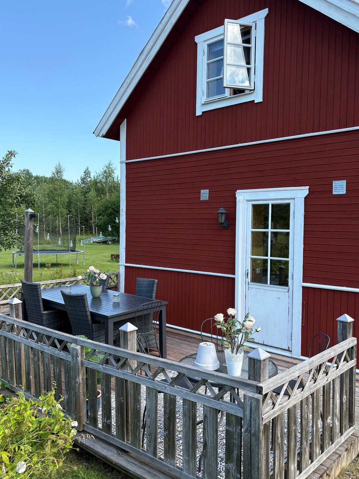 The exterior of the red cabin features a wooden deck with dining furniture, including a rectangular table and several chairs. A small white door leads into the interior, with a large window above it allowing natural light. Lush greenery is visible in the background.