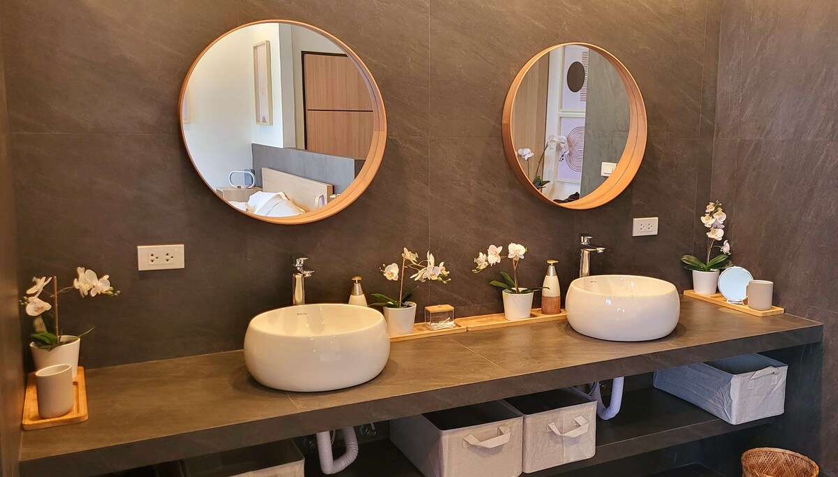 A modern bathroom features two circular mirrors above stylish vessel sinks. White orchids are placed on a bamboo shelf, complementing the minimalist design. Beneath the countertop, storage baskets are neatly arranged, adding functionality to the sophisticated space.