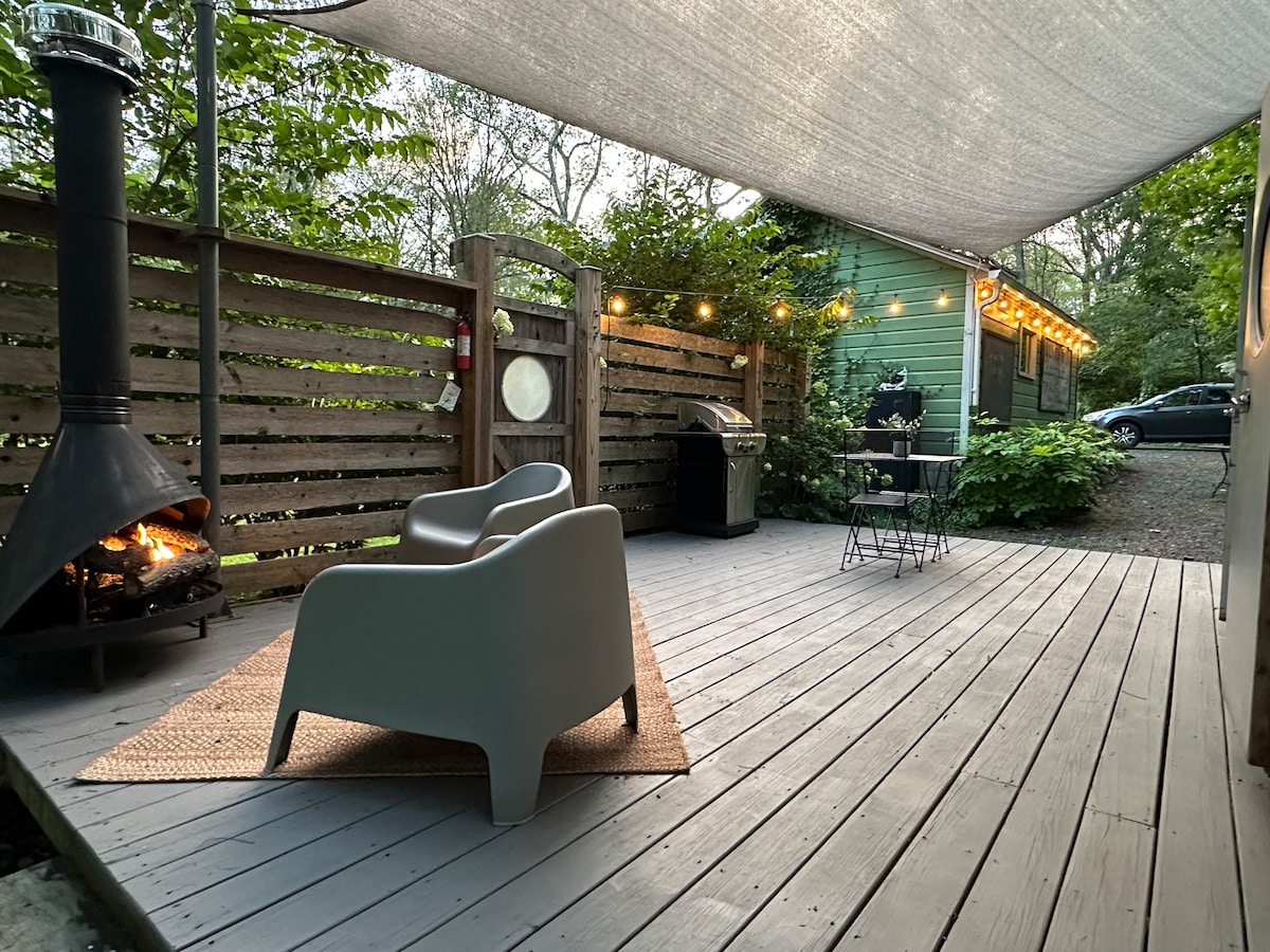 An outdoor deck area features two modern chairs positioned near a firepit, with soft ambient lighting created by string lights overhead. A barbecue grill is visible in the background, alongside a cottage surrounded by greenery, enhancing the semi-secluded atmosphere.