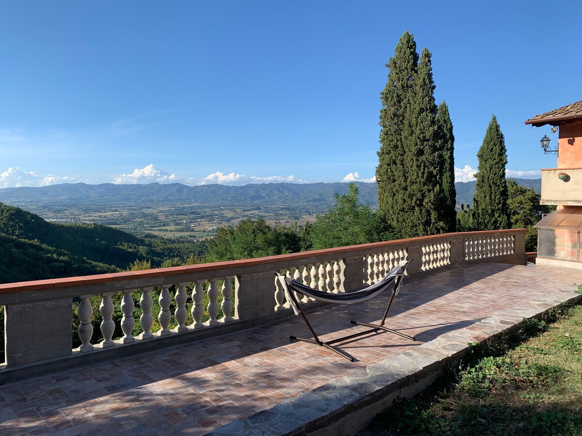 A spacious terrace is viewed, featuring a hammock for relaxation. Lush greenery and rolling hills create a serene backdrop. Cypress trees rise in the distance against a clear blue sky, highlighting the expansive valley below.
