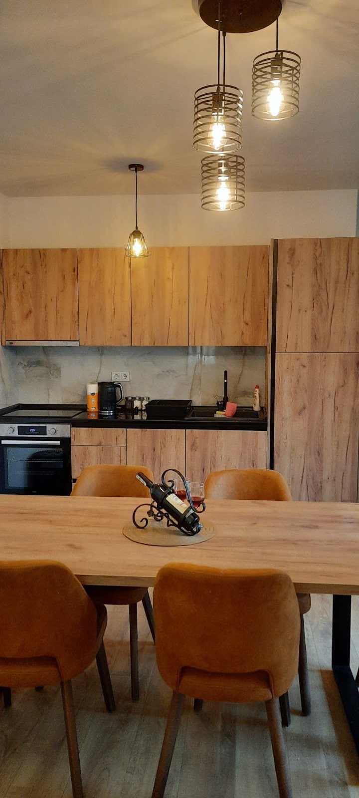 A modern kitchen area features wooden cabinetry with a countertop and a stove. A dining table with four chairs is positioned in the foreground, complemented by decorative lighting fixtures. The space is organized and well-lit, creating a functional and inviting atmosphere.