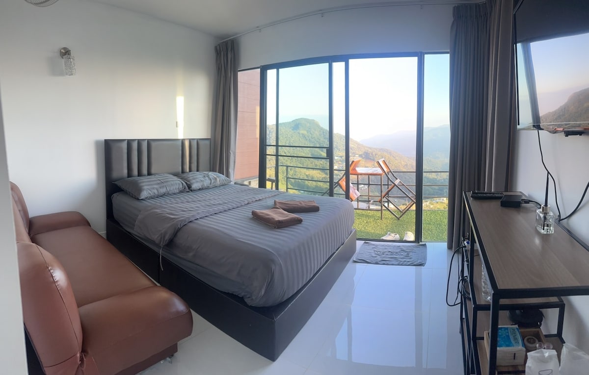 A modern room features a spacious bed with gray linens, complemented by a brown leather sofa. Large glass sliding doors open to reveal a scenic view of mountains, while a flat-screen TV is mounted on the wall and a wood table with two chairs is positioned nearby.