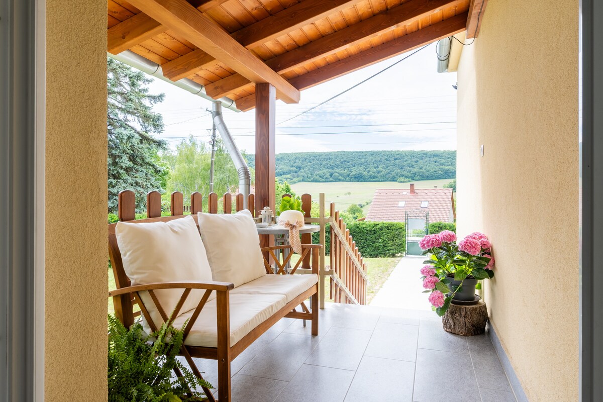 A covered patio is presented, featuring a comfortable seating area with soft cushions. Lush greenery and potted flowers enhance the space. The view offers an open landscape with rolling hills, indicating a peaceful environment. A pathway leads from the patio to the garden area.