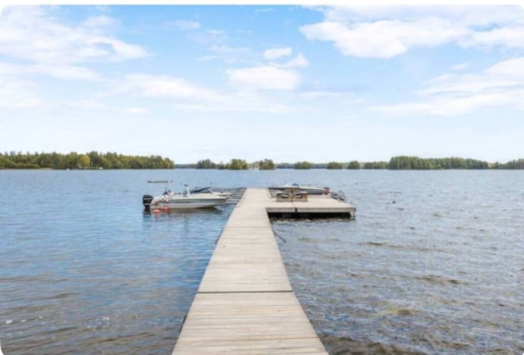 A wooden dock extends into a calm body of water, lined with boats on either side. Lush trees are visible in the background against a clear blue sky, creating a serene environment for leisurely activities.