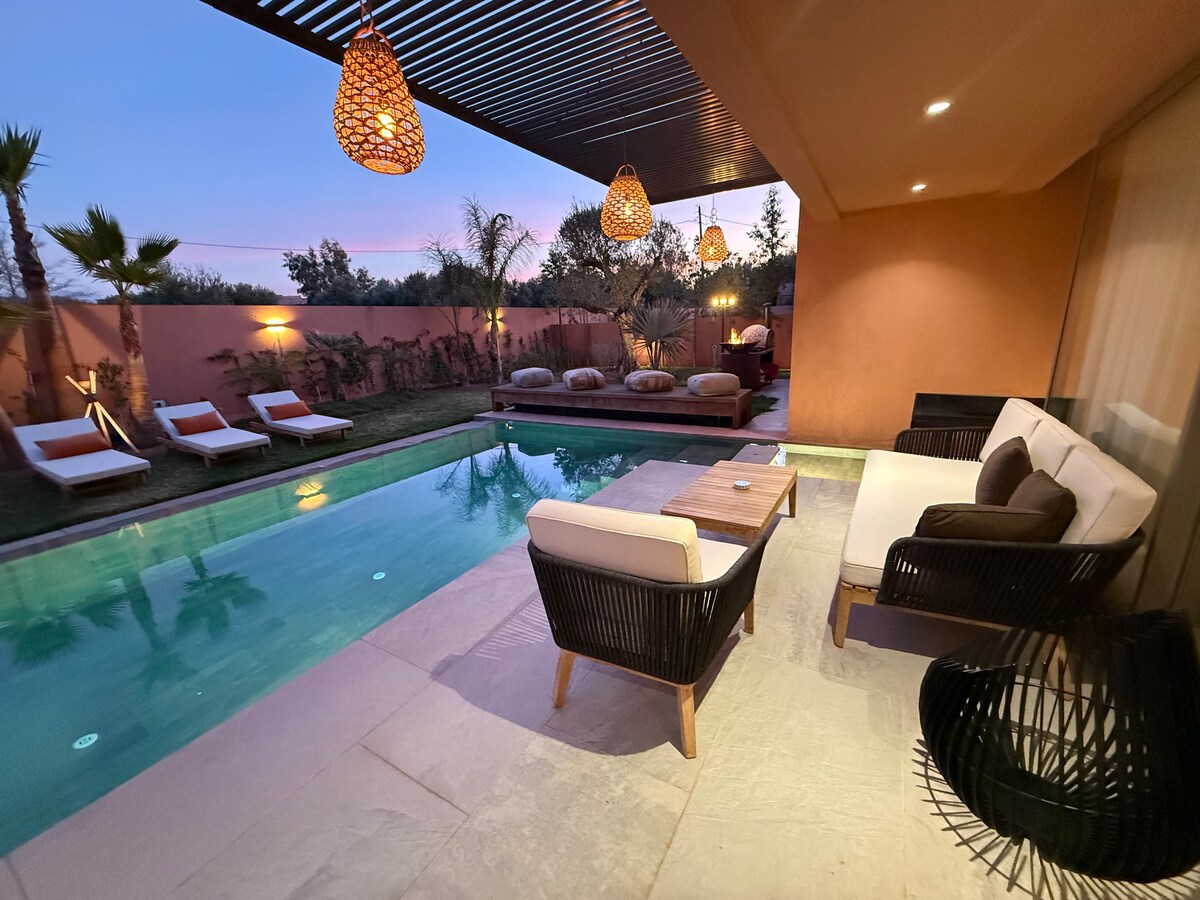 An outdoor lounge area features a comfortable seating arrangement and a wooden table, positioned beside a private heated pool. Soft lounge chairs are placed around the pool, with palm trees providing natural elements. Subtle lighting enhances the atmosphere as twilight descends.