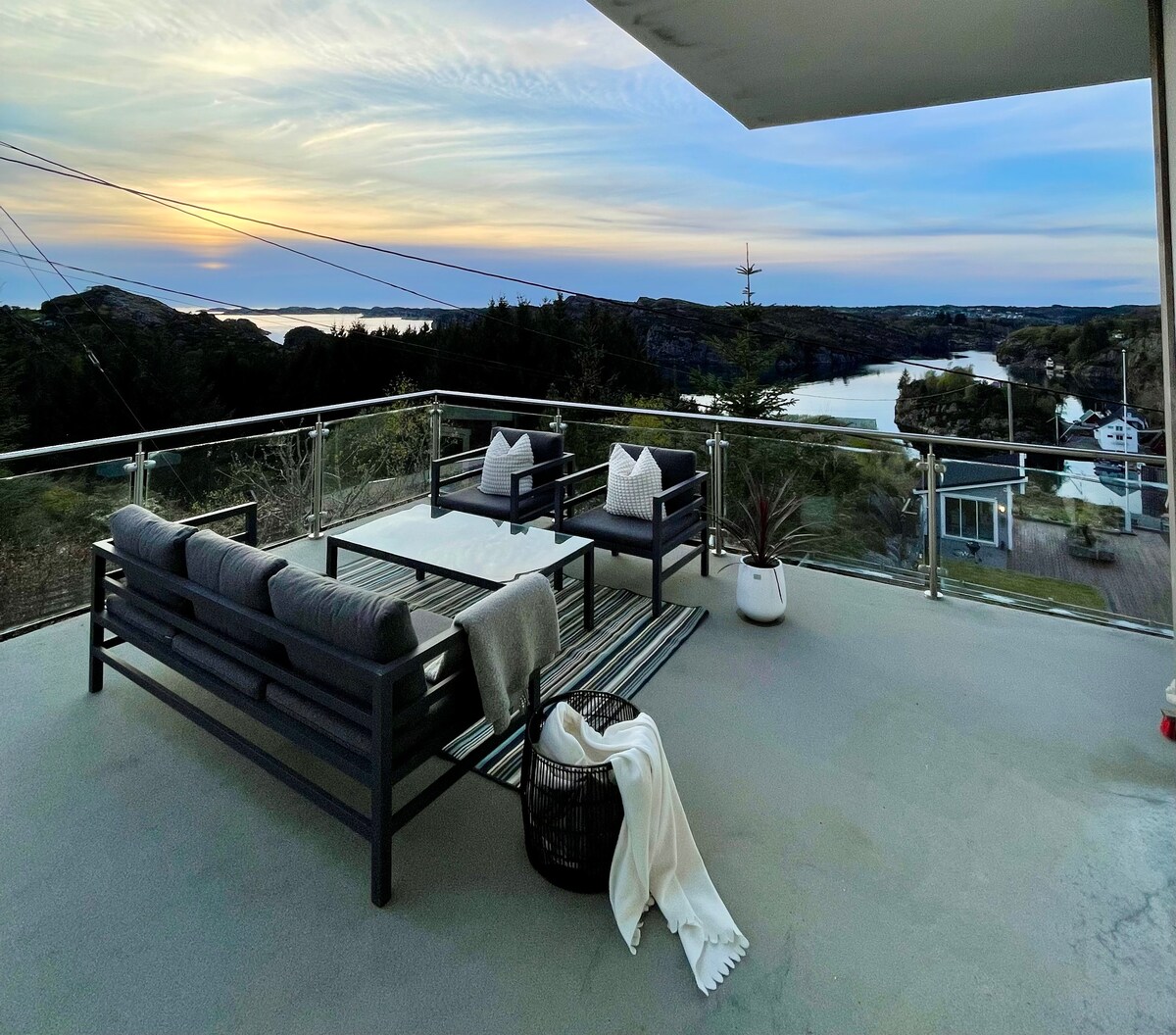 A spacious terrace offers outdoor seating with modern furniture, presenting a scenic view of the coastline and sunset. A cozy throw blanket is draped over a chair, while a potted plant adds a touch of greenery. The sky transitions into soft hues during twilight.