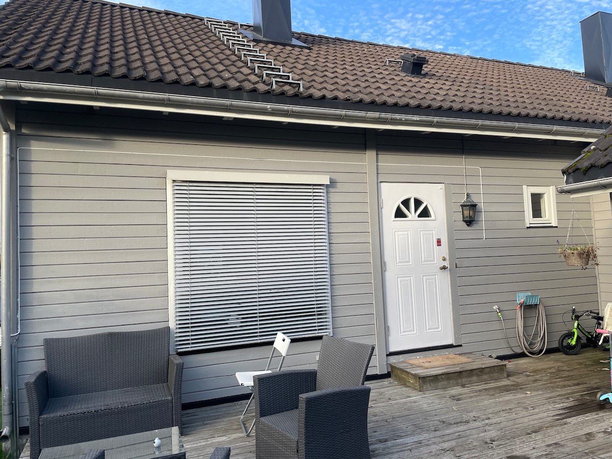 A light gray house exterior features a welcoming front door with a decorative light fixture. Below a large window with white blinds, outdoor seating arrangements made of dark wicker are positioned on a wooden deck, inviting relaxation. A small garden area is visible to the side.