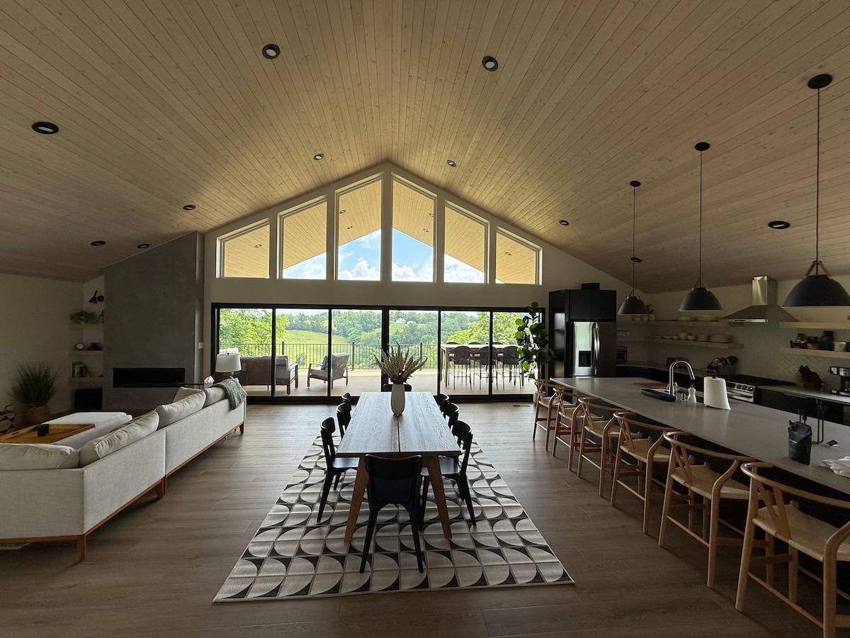 A spacious great room with vaulted ceilings and large windows offers a view of the surrounding countryside. A long dining table with black chairs sits next to a modern kitchen equipped with a dark island. Natural light floods the room, enhancing the open layout.