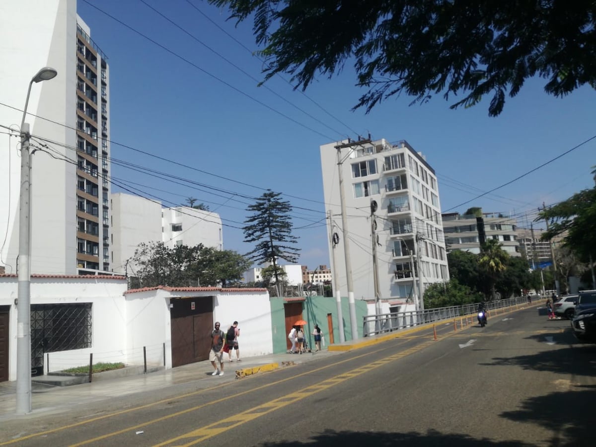 A city street is depicted, lined with modern buildings and trees. Sidewalks are visible where people stroll. Power lines extend across a clear blue sky. The street features a mix of light and shadow, showcasing the area’s urban environment.