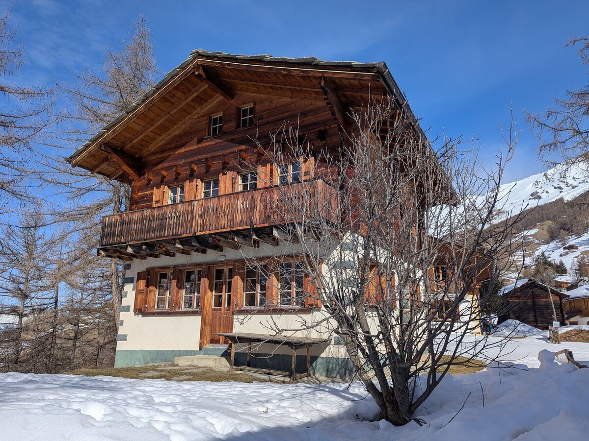 A traditional mountain house is surrounded by snow, featuring a wooden exterior with balconies on the upper floor. Window shutters and multiple doors are visible, along with a bare tree in the foreground. The structure is set against a clear blue sky.