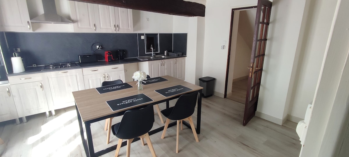 A modern kitchen area is highlighted by a large wooden table with black placemats, surrounded by four sleek black chairs. Light streams in from a nearby window, illuminating the light-colored cabinetry and appliances, including a kettle and coffee maker. A trash bin is discreetly placed in the corner.