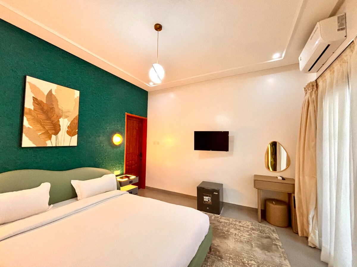 A spacious room features a green accent wall adorned with a decorative wall art piece. A large bed with white linens is positioned prominently. A wall-mounted television is visible, along with a desk and an elegant round mirror. Soft curtains frame a window, offering a calming ambiance.