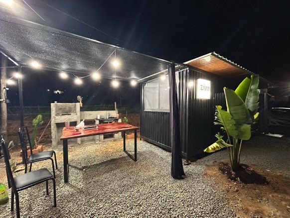 An outdoor dining area is illuminated by string lights, creating a cozy atmosphere. A black table with chairs is set on gravel, surrounded by greenery. The black cabin features a sign and partially transparent curtains that provide a view of the surroundings.