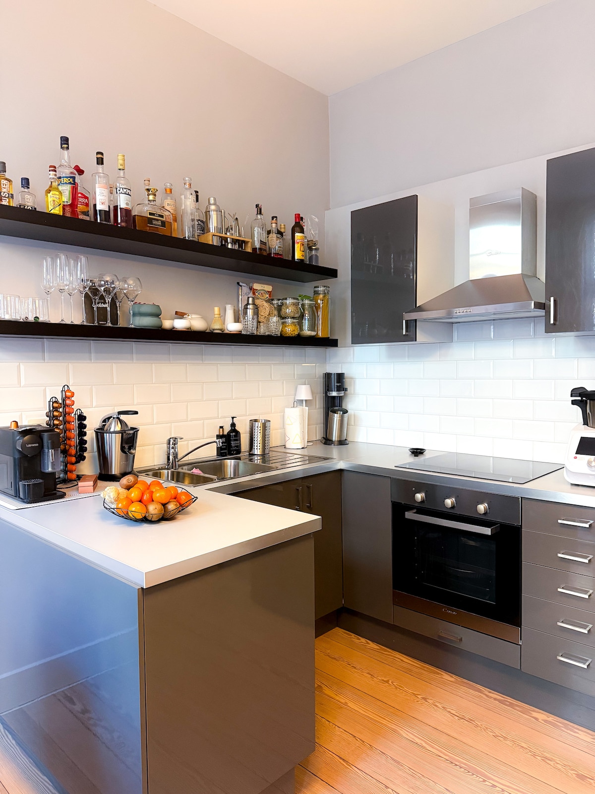 The kitchen features modern cabinetry with a sleek countertop, showcasing a variety of glassware and spirits on open shelves. A bowl of fresh fruit sits on the island, while stainless steel appliances including an oven and a coffee maker are neatly arranged.