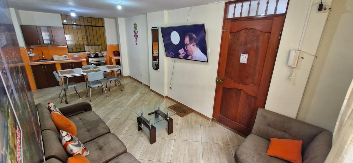 An open living area features a cozy seating arrangement with two comfortable sofas and a glass coffee table. A mounted television is visible on the wall, complemented by a dining space with a table and chairs. Bright orange accents add warmth to the neutral decor.
