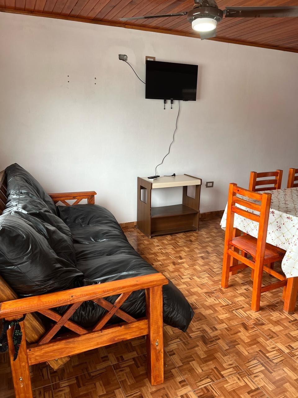 A cozy living area features a dark couch and a wooden coffee table positioned alongside a light wooden dining table with four chairs. A wall-mounted television is visible against a white wall, and light reflects off the patterned flooring.