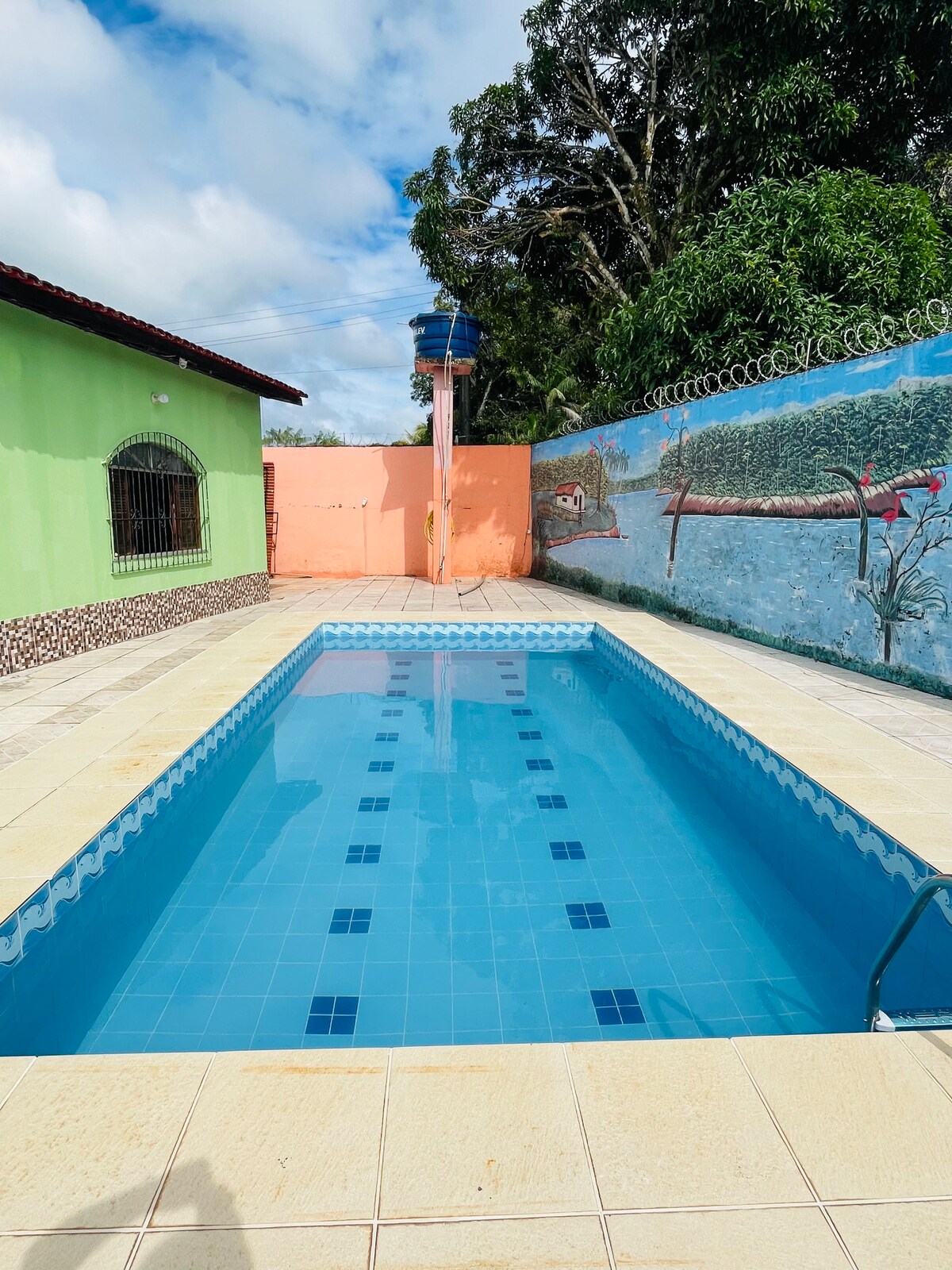 A clear blue swimming pool is set within a spacious outdoor area, surrounded by decorative walls featuring colorful murals. Sunlight reflects off the water, highlighting the pool's clean tiles. Lush greenery is visible in the background, contributing to a serene environment.