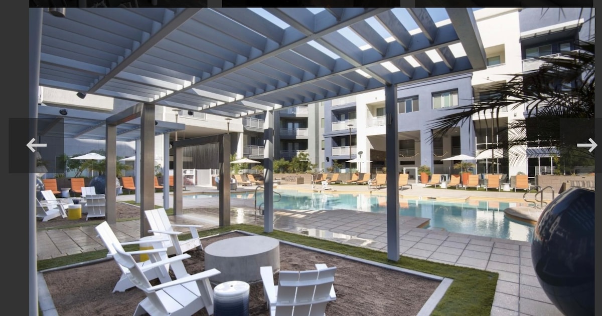 A modern pool area is featured, with a spacious swimming pool surrounded by lounge chairs and umbrellas. White adirondack chairs are arranged around a fire pit, on a patio of laid stone tiles. The building reflects contemporary architecture with large balconies and green landscaping in the background.
