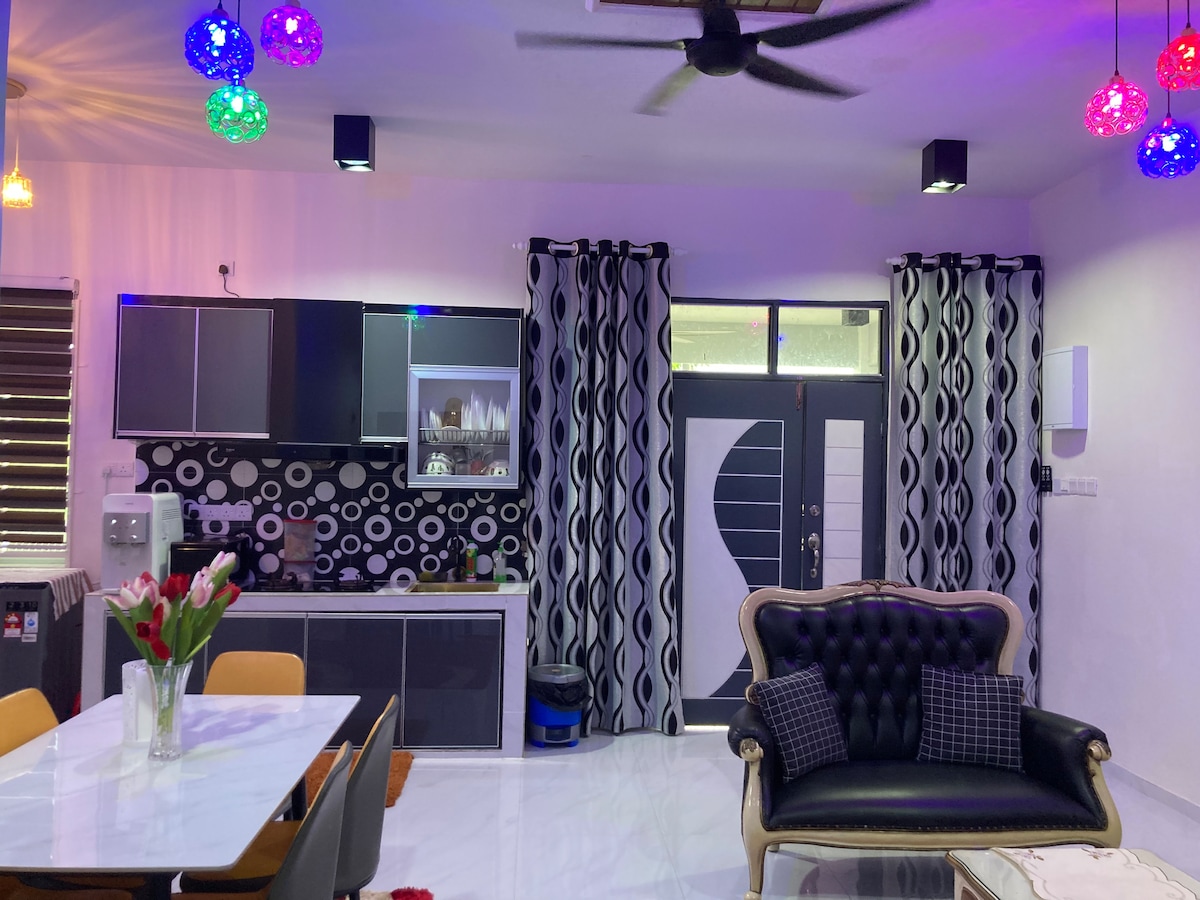 A modern living area is depicted, featuring a dark sofa with decorative cushions and a sleek dining table surrounded by chairs. Colorful pendant lights hang from the ceiling, and a patterned backsplash complements the kitchen area, which is equipped with cabinetry and an open window.