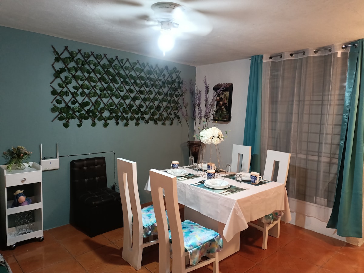 A dining area features a white table set for four, complemented by cushioned chairs. A decorative wall with greenery adds a natural touch. Light gray curtains frame a window, while a ceiling fan provides air circulation.