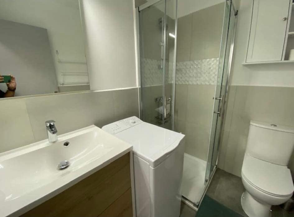 A modern bathroom is equipped with a shower area enclosed by glass. A washing machine is positioned beside a wooden vanity with a sleek sink. Neutral tones create a clean and spacious appearance, complemented by essential fixtures including a toilet and storage shelves.
