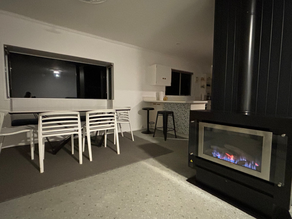 A cozy lounge area highlighted by a gas fireplace featuring a subtle flame display. A dining space with white chairs and a black stool is visible, along with a window offering natural light. The room combines modern amenities with a welcoming layout.