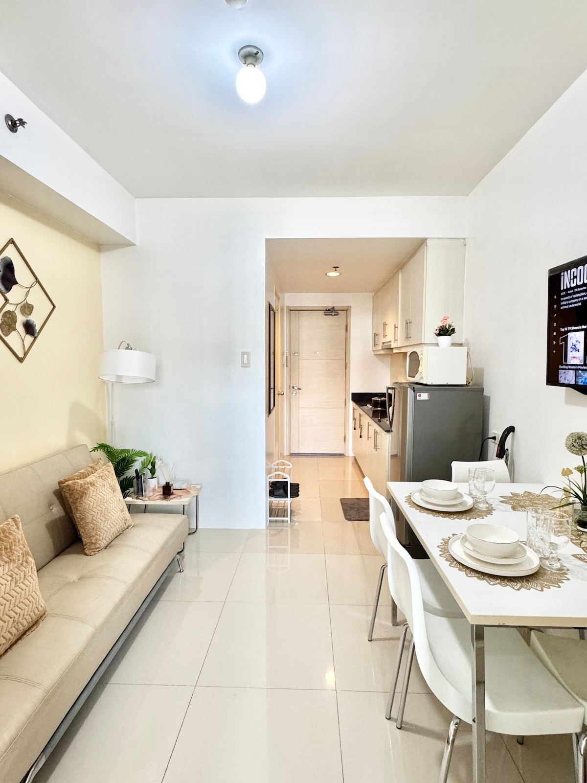 A contemporary living space features a light-toned sofa paired with decorative pillows. A dining area with a table set for four is positioned next to a fully equipped kitchen, with visible appliances including a refrigerator. Bright, neutral-colored walls and polished tile floors enhance the modern aesthetic.