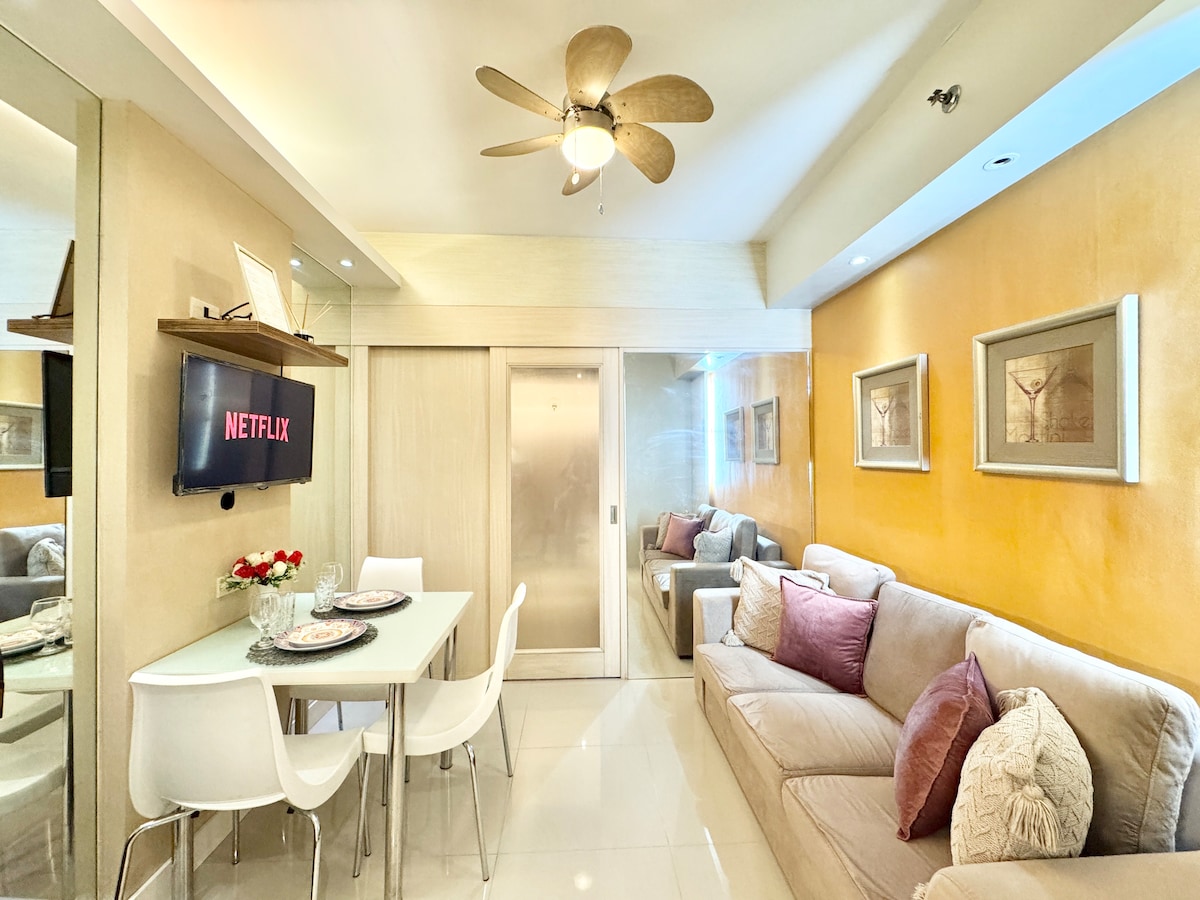 A modern living space is highlighted by warm yellow walls and minimalist decor. A three-seater sofa is paired with a dining table set for two. A television displaying Netflix is mounted on the wall, enhancing the cozy ambiance. Soft lighting adds to the inviting atmosphere.
