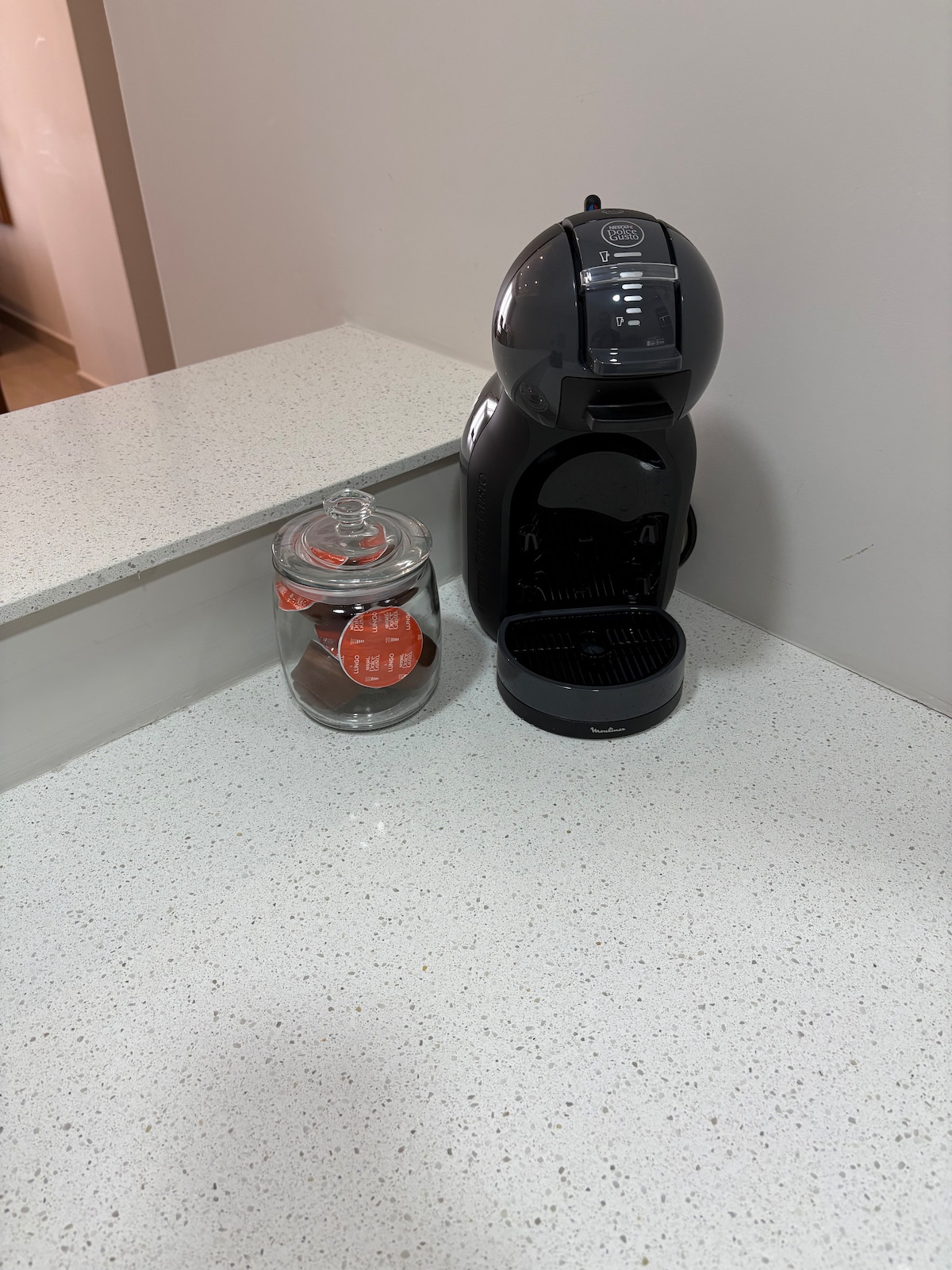 A modern coffee machine sits on a pristine white countertop beside a glass jar containing coffee pods. Soft lighting reflects off the surface, emphasizing the sleek design of the coffee maker.